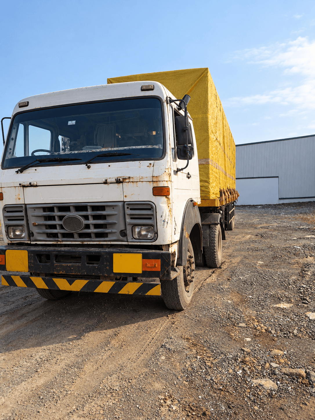 A goods carrier with a yellow tarpaulin over its load, parked beside a warehouse.
