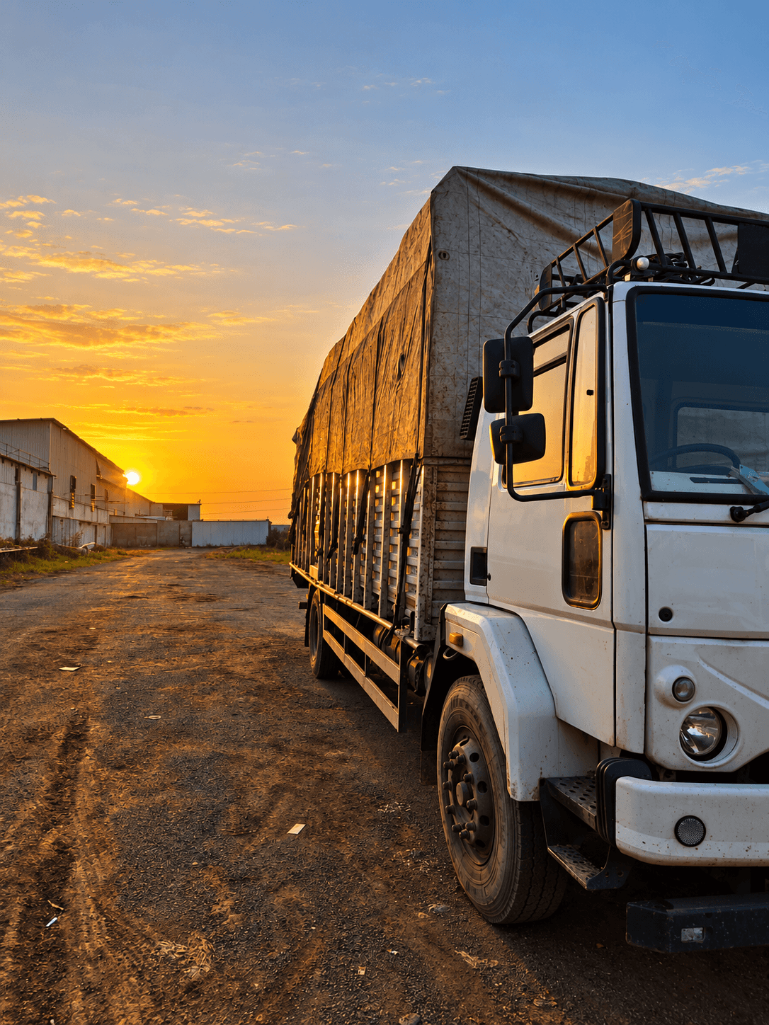 A tarpaulin-covered goods carrier parked in a yard at sunrise, with a warehouse shed behind it.