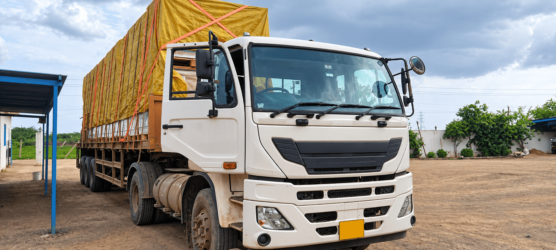 A loaded goods carrier with a yellow tarpaulin over its cargo standing in an industrial yard.